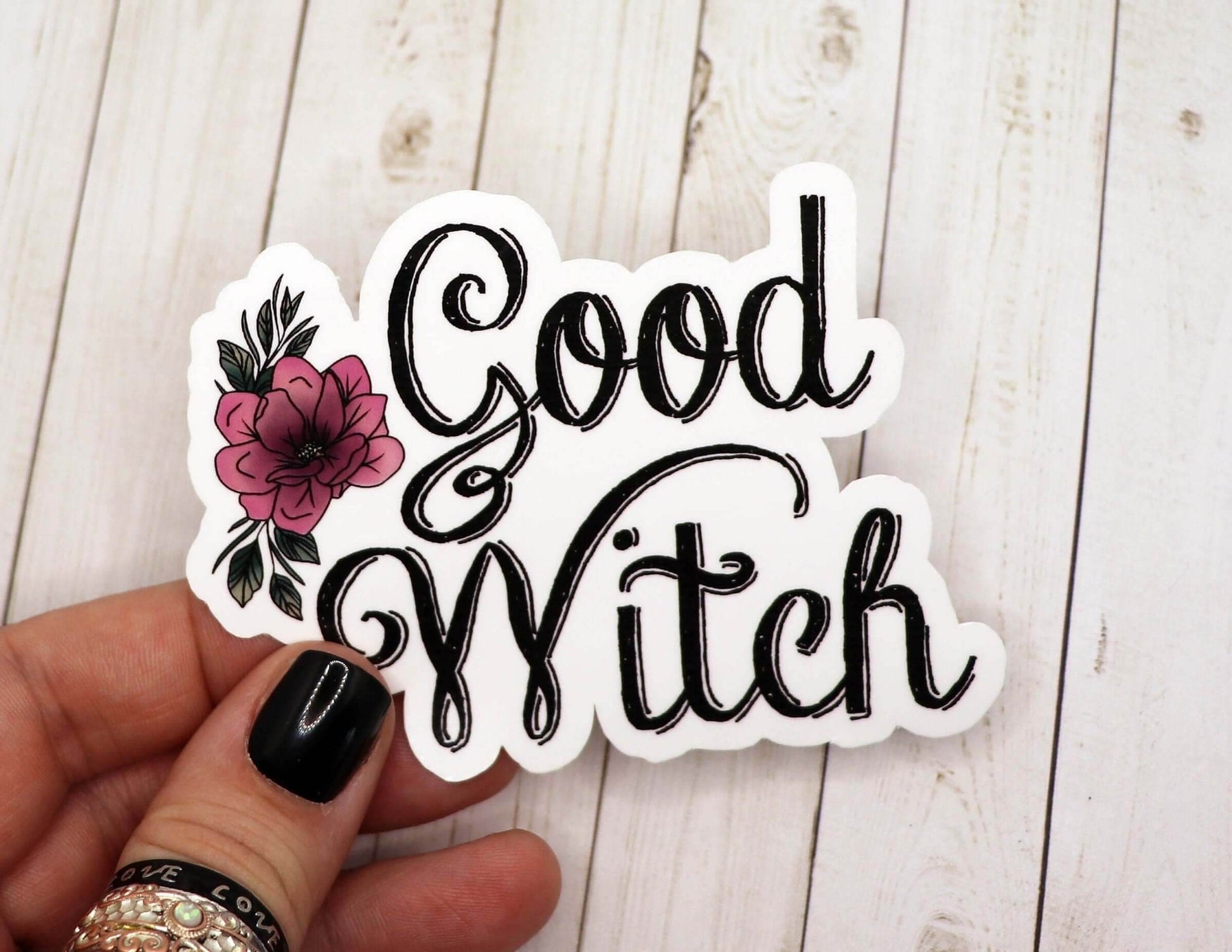 Hand holding a 3-inch Good Witch Sticker with black script and pink flower design on matte vinyl laminate paper against a light wooden background