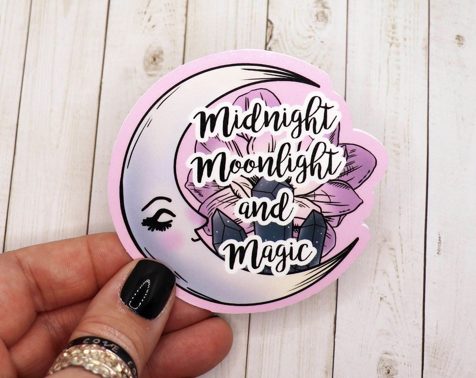 Hand holding a 2.5-inch Midnight Moonlight and Magic vinyl sticker with moon, crystals, and floral design on wooden background