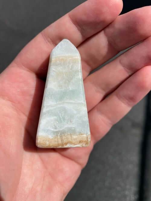 Hand holding a smooth Caribbean Calcite Obelisk crystal with natural blue, green, and yellow hues against a dark background