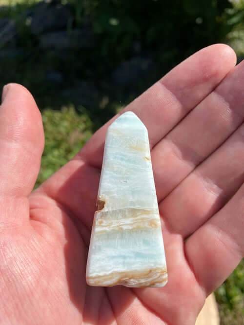 Hand holding a smooth Caribbean Calcite Obelisk crystal with blue, green, and yellow hues against a natural outdoor background.